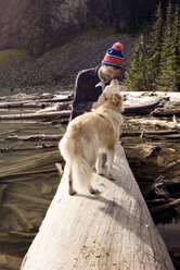 Mann spielt mit Hund auf Baumstamm am Seeufer - CAVF30066