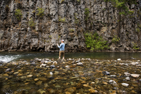 Rückansicht eines Mannes beim Fliegenfischen am Fluss gegen Felsformationen, lizenzfreies Stockfoto