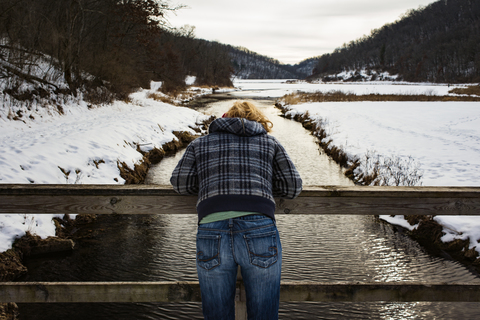 Rückansicht einer Frau, die sich im Winter an ein Geländer über einem Fluss lehnt, lizenzfreies Stockfoto