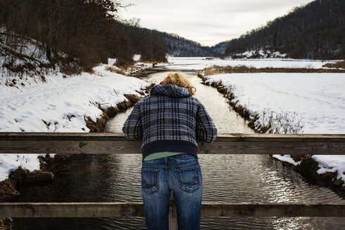 Rückansicht einer Frau, die sich im Winter an ein Geländer über einem Fluss lehnt - CAVF30141