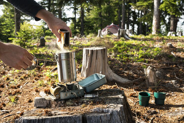 Ausgeschnittenes Bild eines Mannes, der Essen in einen Behälter über einem Campingkocher im Wald gießt - CAVF30190