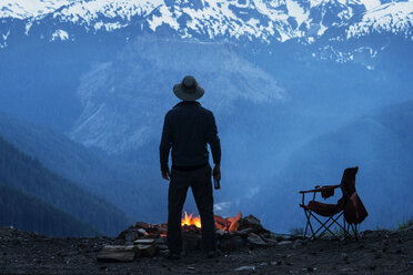 Man standing by campfire against snowcapped mountains - CAVF30195