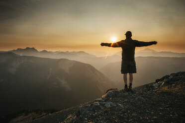 Rear view of man standing with arms outstretched on mountain against sky during sunset - CAVF30222