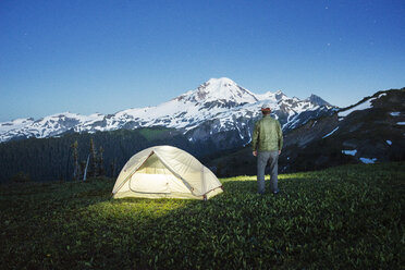 Rear view of man standing by illuminated tent on grassy field against mountain at dusk - CAVF30289