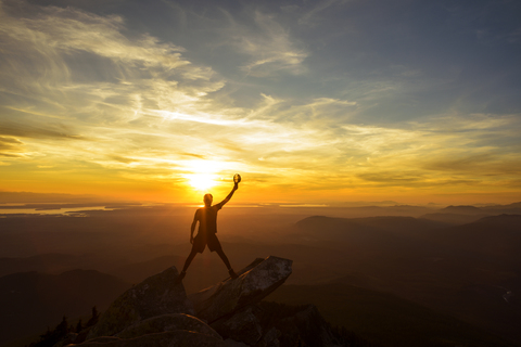 Silhouette eines Wanderers, der eine Mütze mit erhobener Hand hält, während er bei Sonnenuntergang auf dem Gipfel eines Berges steht, lizenzfreies Stockfoto