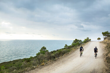 Rear view of male friends cycling on road by sea against cloudy sky - CAVF30466