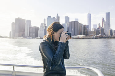 Side view of woman photographing while standing on boat against cityscape - CAVF30491