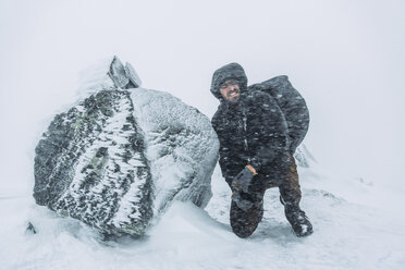Junger Rucksacktourist kniet während eines Schneesturms neben einem Felsen - FOLF02520