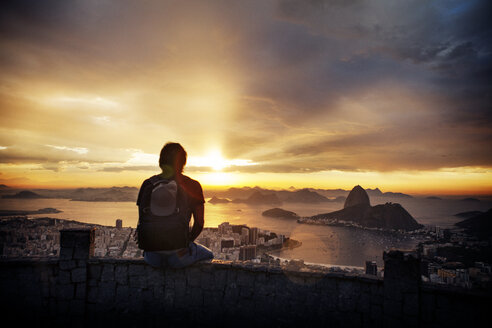 Rear view of man sitting on retaining wall at observation point against city and cloudy sky - CAVF30603