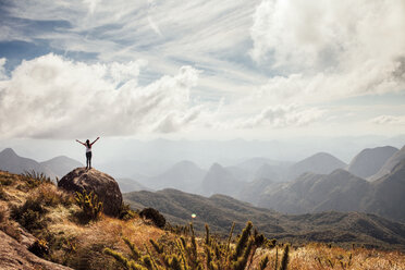 Fernblick auf eine Wanderin, die mit ausgestreckten Armen auf einem Berg vor bewölktem Himmel steht - CAVF30617