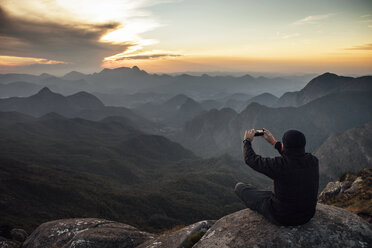 Rückansicht eines Wanderers, der während des Sonnenuntergangs auf einem Berg sitzt und mit seinem Smartphone fotografiert - CAVF30619