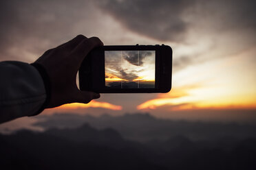 Ausgeschnittenes Bild eines Mannes, der mit seinem Smartphone bei Sonnenuntergang den bewölkten Himmel fotografiert - CAVF30620