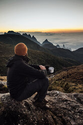 Side view of man holding mug while sitting on mountain during sunset - CAVF30621