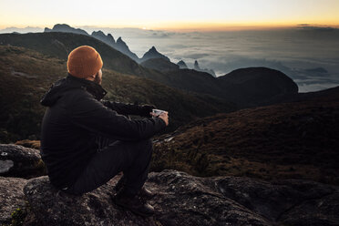 Man holding mug while sitting on mountain during sunset - CAVF30622