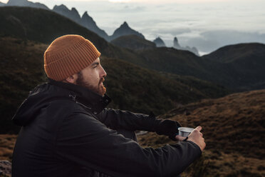 Thoughtful man holding mug while sitting on mountain - CAVF30623