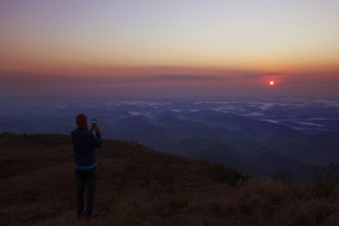 Mann fotografiert auf einem Berg stehend den Sonnenuntergang - CAVF30634