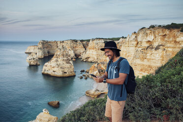 Lächelnder Mann, der ein Telefon benutzt, während er auf einer Klippe am Meer steht - CAVF30645