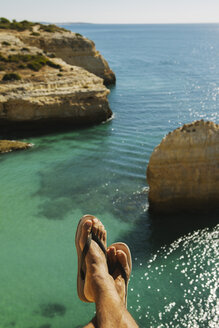 Low section of man wearing slippers resting on cliff against sea - CAVF30655