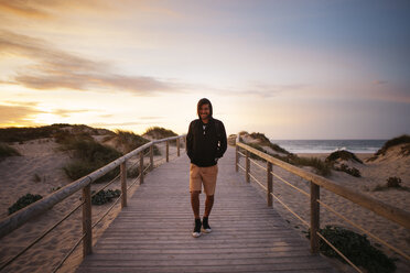 Smiling hiker walking on footbridge at beach against sky during sunset - CAVF30666