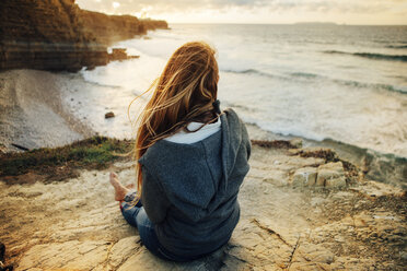 Rear view of woman sitting on cliff and looking at view during sunset - CAVF30676