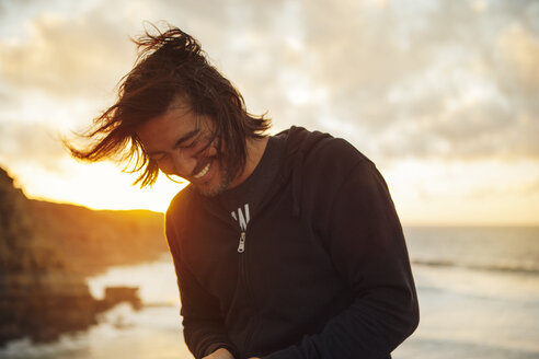Glücklicher Mann lächelnd am Strand stehend gegen bewölkten Himmel bei Sonnenuntergang - CAVF30677