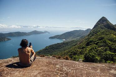 Rear view of man photographing sea while sitting on mountain against sky - CAVF30713