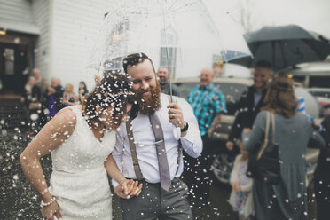 Glückliches frisch vermähltes Paar, das mit einem Regenschirm spazieren geht, während die Leute im Hintergrund während der Hochzeitszeremonie stehen - CAVF30846