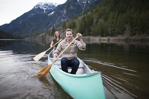 Glückliches junges Paar beim Kanufahren auf einem See vor den Bergen - CAVF30869