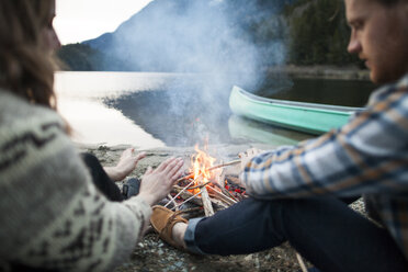 Young couple warming hands by campfire at lakeshore - CAVF30875