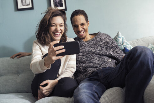 Junges Paar sitzt auf dem Sofa, schaut auf sein Smartphone und lacht - FOLF03327