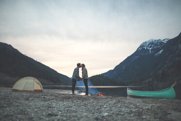 Junges Paar hält sich in voller Länge an den Händen und küsst sich in der Abenddämmerung am Seeufer vor den Bergen - CAVF30879