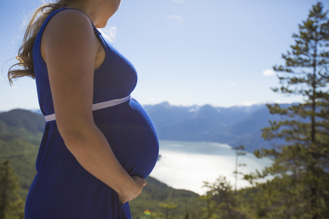 Mittelteil einer schwangeren Frau vor Bergen und Himmel, lizenzfreies Stockfoto