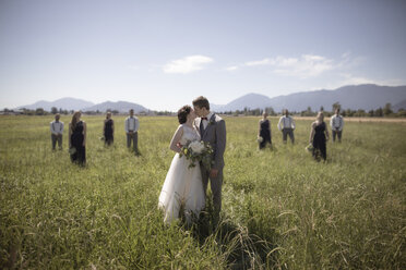 Newlywed Paar küssen, während stehend auf grasbewachsenen Feld mit groomsmen und Brautjungfern im Hintergrund während sonnigen Tag - CAVF30890