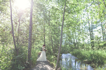 Seitenansicht der Braut, die einen Blumenstrauß hält, während sie inmitten von Bäumen im Wald steht - CAVF30897