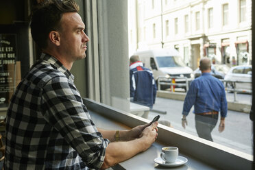 Mann mit Handy in der Hand und Blick durch das Fenster eines Cafés - FOLF03715