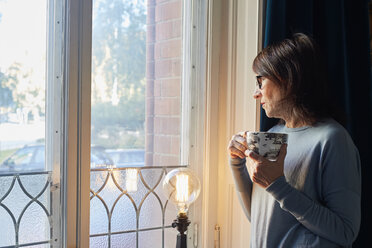 Frau schaut durch ein Fenster und hält eine Kaffeetasse - FOLF03734