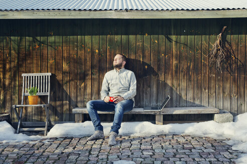 Mittlerer erwachsener Mann sitzt auf einer Bank neben einem Holzschuppen - FOLF03841