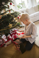 Kleiner blonder Junge überprüft Weihnachtsgeschenke auf dem Boden sitzend - FOLF03903