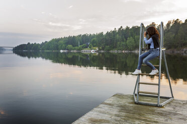 Woman sitting on ladder, photographing lake at Vato, Sweden - FOLF04351