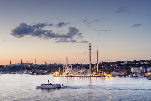 Beleuchteter Vergnügungspark Grona Lund in Stockholm bei Sonnenuntergang, über das Wasser gesehen, mit vorbeifahrendem Schiff - FOLF04390