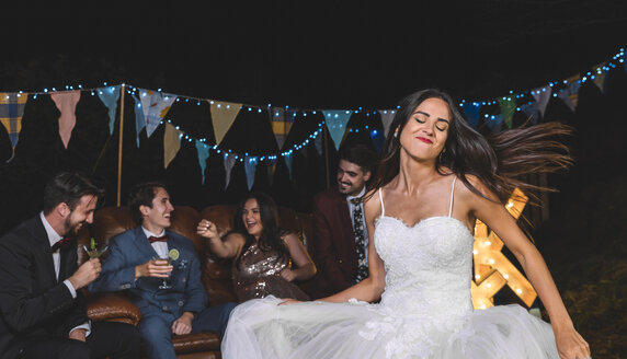 Happy bride dancing on a night field party with groom and friends in the background - DAPF00936