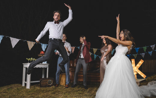 Surprised bride looking at man jumping on a night field party with friends - DAPF00942