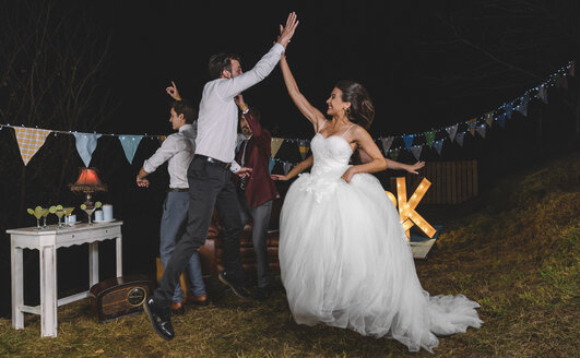 Happy bride jumping and giving high five with young man on a night field party with friends - DAPF00943