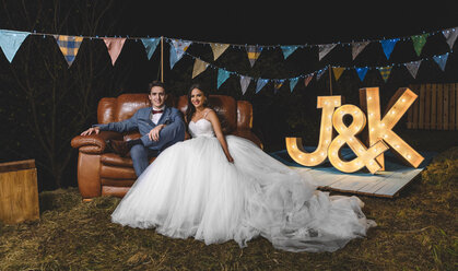 Portrait of happy wedding couple sitting on sofa on a night field party - DAPF00948