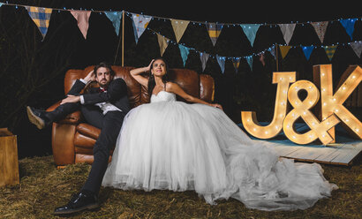 Portrait of wedding couple posing sitting on sofa on a night field party - DAPF00950