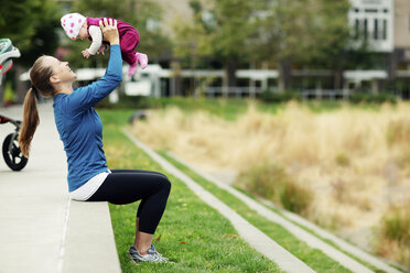 Side view of woman lifting baby while sitting on retaining wall at park - CAVF31162