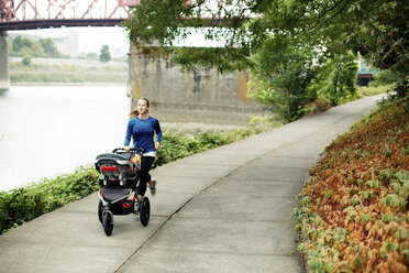 Frau joggt mit Kinderwagen auf dem Fußweg am Fluss - CAVF31165