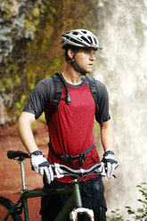 Mountainbiker hält Fahrrad und steht gegen Wasserfall - CAVF31234