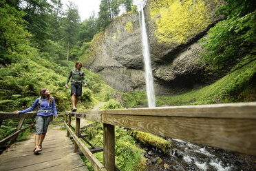 Paar, das auf einem Steg bei einem Wasserfall im Wald spazieren geht - CAVF31255