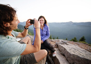 Woman looking at boyfriend photographing while sitting on mountain - CAVF31270